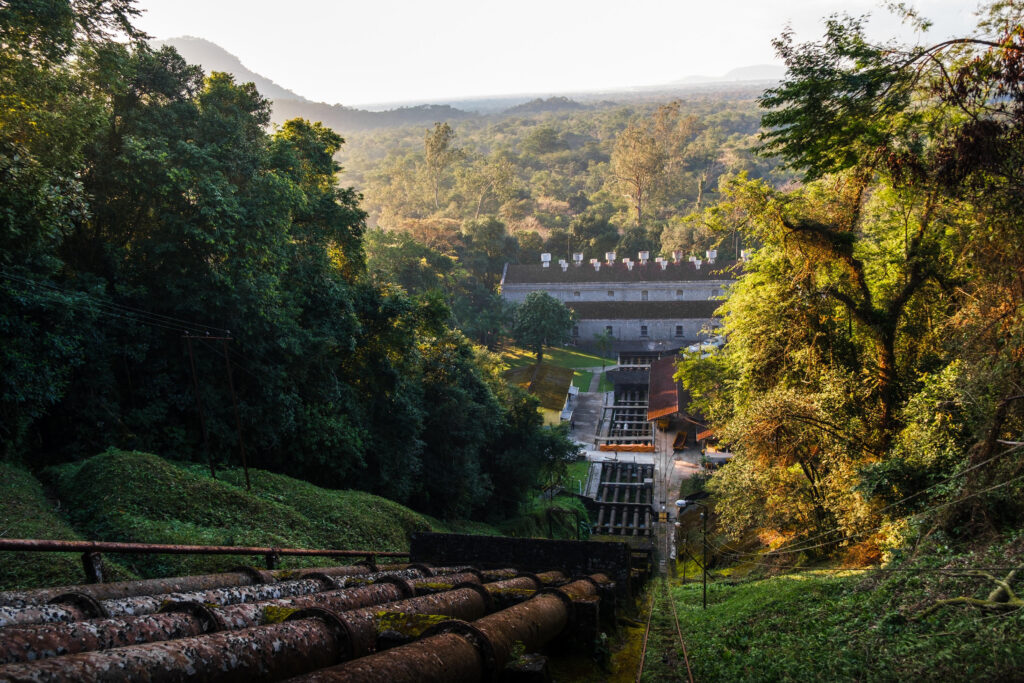 Uma foto aérea de uma usina hidrelétrica em uma encosta íngreme, com grandes tubulações de aço oxidado (penstocks) descendo a colina densamente florestada à esquerda. À direita, trilhos de um pequeno sistema de transporte sobem a encosta. No complexo industrial na base da colina, há um grande edifício de telhado de duas águas com múltiplos tubos de ventilação, além de outros edifícios menores e mais infraestrutura. A usina está cercada por uma vasta paisagem de floresta tropical densa que se estende até o horizonte sob a luz quente e difusa do pôr do sol. A imagem captura o contraste entre a infraestrutura industrial e a natureza circundante.