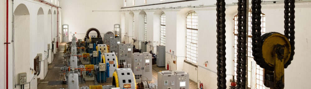 Uma vista aérea elevada do interior de um grande salão de maquinário em uma usina hidrelétrica. Fileiras ordenadas de equipamentos industriais pesados, incluindo múltiplas unidades geradoras de energia com grandes turbinas cinzentas e geradores com carcaças amarelas brilhantes, estão dispostas no centro. Elas são flanqueadas por altos painéis de controle cinzentos e armários elétricos. O salão tem um alto teto de metal treliçado exposto com clarabóias e luminárias pendentes, e as paredes brancas são pontuadas por grandes janelas arqueadas com grades de segurança. O chão tem um padrão de azulejos geométricos desgastados. À direita, um grande gancho de guindaste e cadeias de carga pendem de uma estrutura superior. Tubos de incêndio vermelhos e outros equipamentos menores são visíveis ao longo das paredes. Esta imagem captura a complexidade e a escala do maquinário no complexo da usina hidrelétrica do Porto de Santos.