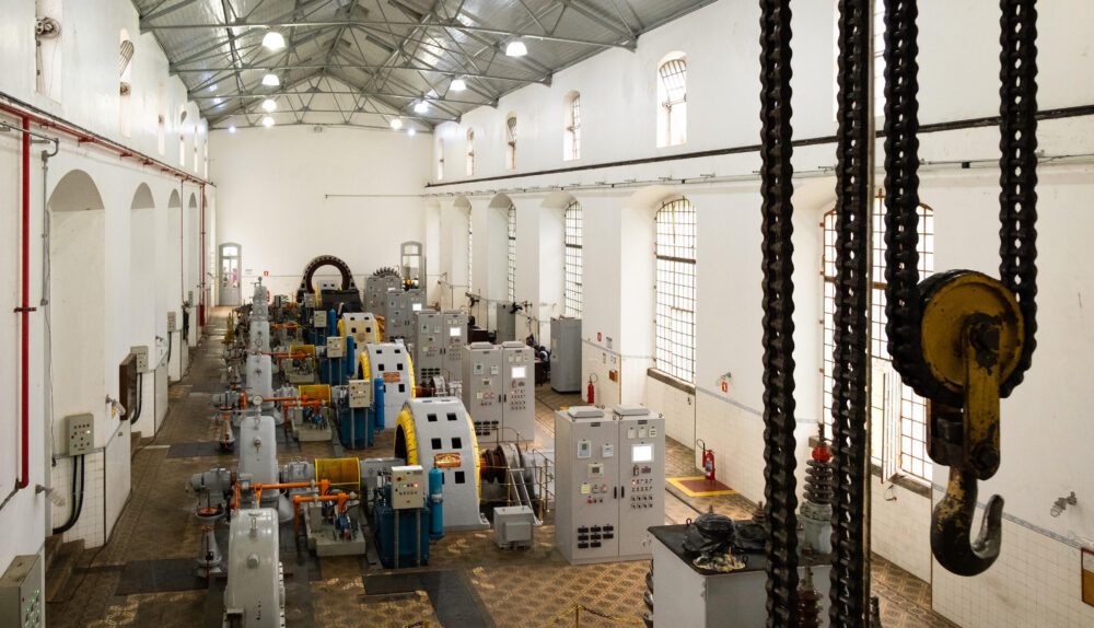 Uma vista aérea elevada do interior de um grande salão de maquinário em uma usina hidrelétrica. Fileiras ordenadas de equipamentos industriais pesados, incluindo múltiplas unidades geradoras de energia com grandes turbinas cinzentas e geradores com carcaças amarelas brilhantes, estão dispostas no centro. Elas são flanqueadas por altos painéis de controle cinzentos e armários elétricos. O salão tem um alto teto de metal treliçado exposto com clarabóias e luminárias pendentes, e as paredes brancas são pontuadas por grandes janelas arqueadas com grades de segurança. O chão tem um padrão de azulejos geométricos desgastados. À direita, um grande gancho de guindaste e cadeias de carga pendem de uma estrutura superior. Tubos de incêndio vermelhos e outros equipamentos menores são visíveis ao longo das paredes. Esta imagem captura a complexidade e a escala do maquinário no complexo da usina hidrelétrica do Porto de Santos.