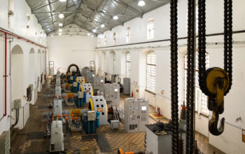 Uma vista aérea elevada do interior de um grande salão de maquinário em uma usina hidrelétrica. Fileiras ordenadas de equipamentos industriais pesados, incluindo múltiplas unidades geradoras de energia com grandes turbinas cinzentas e geradores com carcaças amarelas brilhantes, estão dispostas no centro. Elas são flanqueadas por altos painéis de controle cinzentos e armários elétricos. O salão tem um alto teto de metal treliçado exposto com clarabóias e luminárias pendentes, e as paredes brancas são pontuadas por grandes janelas arqueadas com grades de segurança. O chão tem um padrão de azulejos geométricos desgastados. À direita, um grande gancho de guindaste e cadeias de carga pendem de uma estrutura superior. Tubos de incêndio vermelhos e outros equipamentos menores são visíveis ao longo das paredes. Esta imagem captura a complexidade e a escala do maquinário no complexo da usina hidrelétrica do Porto de Santos.