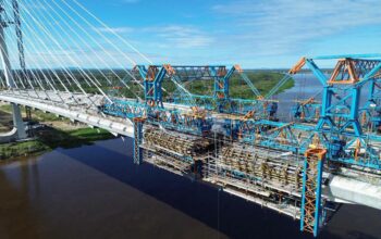 Vista aérea de drone da ponte internacional da Rota Bioceânica em construção sobre um rio, mostrando as complexas estruturas metálicas que se preparam para o "beijo das aduelas", a conexão final entre Brasil e Paraguai.