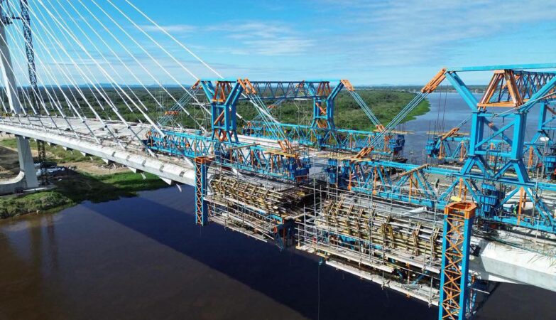 Vista aérea de drone da ponte internacional da Rota Bioceânica em construção sobre um rio, mostrando as complexas estruturas metálicas que se preparam para o "beijo das aduelas", a conexão final entre Brasil e Paraguai.