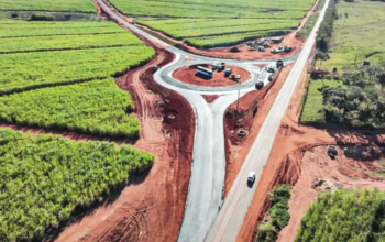 Uma vista aérea de drone de uma rotatória de tráfego em construção e recém-pavimentada, situada em uma paisagem rural rodeada por densos campos de cana-de-açúcar e áreas de terra vermelha exposta. Vários caminhões de construção e maquinários estão visíveis dentro do círculo central da rotatória e em um local de trabalho adjacente, com um único carro branco circulando em uma estrada de acesso. Esta imagem exemplifica o tipo de projeto de infraestrutura coberto pela iniciativa