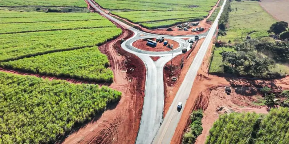 Uma vista aérea de drone de uma rotatória de tráfego em construção e recém-pavimentada, situada em uma paisagem rural rodeada por densos campos de cana-de-açúcar e áreas de terra vermelha exposta. Vários caminhões de construção e maquinários estão visíveis dentro do círculo central da rotatória e em um local de trabalho adjacente, com um único carro branco circulando em uma estrada de acesso. Esta imagem exemplifica o tipo de projeto de infraestrutura coberto pela iniciativa