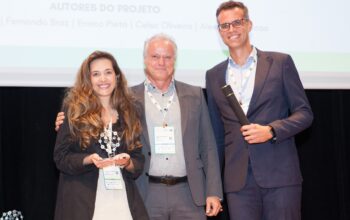 Uma fotografia de premiação num palco, mostrando (da esquerda para a direita) Maria Gabriela da Climb Consulting, segurando um troféu transparente com detalhes prateados, ao lado de Sergei Fortes do Sinaenco, que está no centro com o braço no ombro dela, e Bernardo Etges da Climb Consulting, segurando um rolo de certificado e sorrindo. Os três profissionais estão formalmente vestidos, usando crachás de identificação e posando para a câmera.