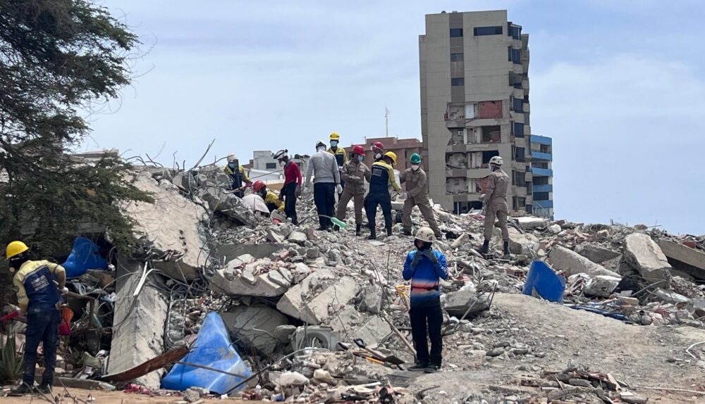 Uma cena panorâmica mostra os escombros de um desastre, provavelmente as consequências de um terremoto na Venezuela, com uma equipe de busca e salvamento trabalhando ativamente sobre uma pilha maciça de destroços de concreto. Vários socorristas, usando capacetes de proteção em cores variadas (como amarelo, vermelho, verde e branco) e fardas de trabalho, estão no topo da pilha, colaborando para remover fragmentos e procurar sobreviventes. Pelo menos sete pessoas estão visíveis no topo, com poses que sugerem trabalho em equipe, incluindo uma figura em primeiro plano, no nível do chão, olhando para o local com as mãos perto da boca. Um prédio alto, com danos visíveis nas paredes externas e janelas quebradas, está de pé ao fundo, contrastando com o nível de destruição no chão. O chão à frente é de terra batida com detritos soltos e galhos. O céu está parcialmente nublado. A composição foca na interação humana com a escala dos escombros.