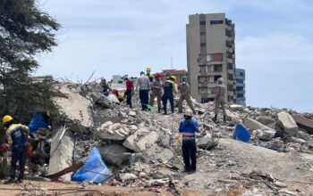 Uma cena panorâmica mostra os escombros de um desastre, provavelmente as consequências de um terremoto na Venezuela, com uma equipe de busca e salvamento trabalhando ativamente sobre uma pilha maciça de destroços de concreto. Vários socorristas, usando capacetes de proteção em cores variadas (como amarelo, vermelho, verde e branco) e fardas de trabalho, estão no topo da pilha, colaborando para remover fragmentos e procurar sobreviventes. Pelo menos sete pessoas estão visíveis no topo, com poses que sugerem trabalho em equipe, incluindo uma figura em primeiro plano, no nível do chão, olhando para o local com as mãos perto da boca. Um prédio alto, com danos visíveis nas paredes externas e janelas quebradas, está de pé ao fundo, contrastando com o nível de destruição no chão. O chão à frente é de terra batida com detritos soltos e galhos. O céu está parcialmente nublado. A composição foca na interação humana com a escala dos escombros.