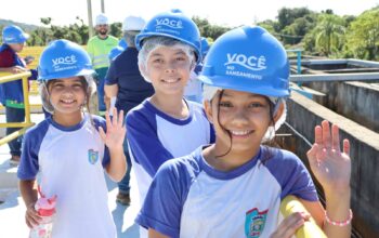 Uma fotografia colorida em plano médio captura três crianças sorridentes em primeiro plano, usando uniformes escolares brancos e azuis, toucas de proteção e capacetes de segurança azuis com a inscrição "VOCÊ NO SANEAMENTO" durante uma visita guiada educativa em uma instalação de tratamento da Iguá Saneamento. Duas das crianças, que estão no centro e à direita, acenam alegremente para a câmera. Elas estão sobre uma passarela de concreto e metal, com grandes tanques de tratamento de concreto visíveis ao fundo e vegetação densa sob um céu claro e ensolarado. Outros estudantes e adultos (um guia de colete refletivo) também usando capacetes são vistos ao fundo. A criança da esquerda segura uma garrafa de água infantil rosa. Esta imagem captura uma iniciativa educativa da Iguá Saneamento.