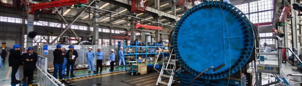 Uma visão detalhada e ampla do interior de uma movimentada fábrica industrial de fabricação pesada. À direita, destaca-se um componente industrial maciço, cilíndrico e azul brilhante, com uma enorme flange repleta de dezenas de parafusos, posicionado em um suporte azul. Este equipamento robusto sugere a fabricação de componentes críticos para o setor de energias renováveis, como partes para turbinas hidrelétricas ou eólicas de grande porte. Um trabalhador com capacete branco e macacão azul está no topo do equipamento, ajustando conexões perto de manômetros e mangueiras. Uma escada de metal se apoia na base da estrutura. À esquerda, um grupo de oito pessoas, incluindo engenheiros e inspetores vestindo capacetes azuis e brancos e casacos azuis, está reunido no piso de concreto, alguns observando pranchetas ou o equipamento. O vasto espaço da oficina é caracterizado por sua estrutura metálica, guindastes de pórtico vermelhos suspensos no teto alto, grandes janelas que deixam entrar luz natural e várias outras máquinas e prateleiras de estoque ao fundo. Placas informativas e cercas de segurança também são visíveis.