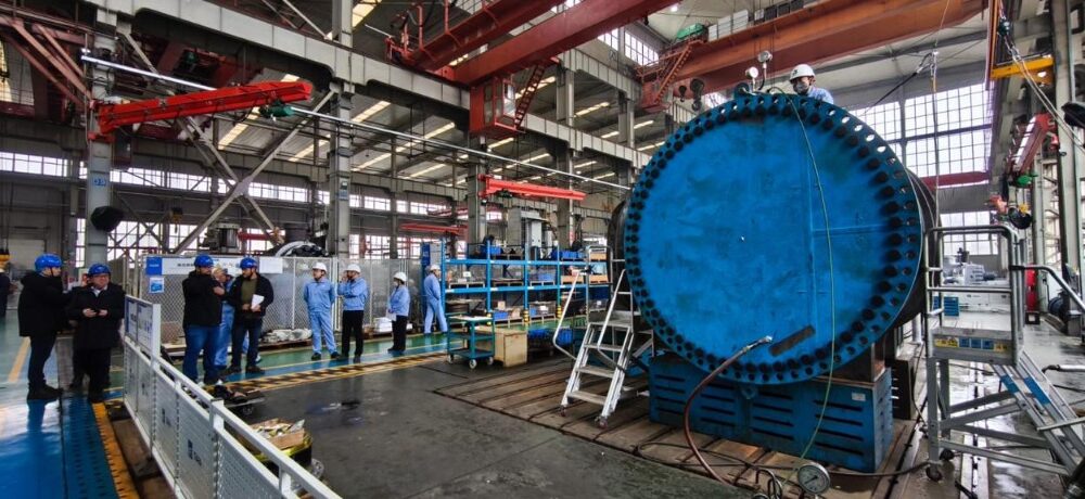 Uma visão detalhada e ampla do interior de uma movimentada fábrica industrial de fabricação pesada. À direita, destaca-se um componente industrial maciço, cilíndrico e azul brilhante, com uma enorme flange repleta de dezenas de parafusos, posicionado em um suporte azul. Este equipamento robusto sugere a fabricação de componentes críticos para o setor de energias renováveis, como partes para turbinas hidrelétricas ou eólicas de grande porte. Um trabalhador com capacete branco e macacão azul está no topo do equipamento, ajustando conexões perto de manômetros e mangueiras. Uma escada de metal se apoia na base da estrutura. À esquerda, um grupo de oito pessoas, incluindo engenheiros e inspetores vestindo capacetes azuis e brancos e casacos azuis, está reunido no piso de concreto, alguns observando pranchetas ou o equipamento. O vasto espaço da oficina é caracterizado por sua estrutura metálica, guindastes de pórtico vermelhos suspensos no teto alto, grandes janelas que deixam entrar luz natural e várias outras máquinas e prateleiras de estoque ao fundo. Placas informativas e cercas de segurança também são visíveis.