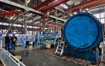 Uma visão detalhada e ampla do interior de uma movimentada fábrica industrial de fabricação pesada. À direita, destaca-se um componente industrial maciço, cilíndrico e azul brilhante, com uma enorme flange repleta de dezenas de parafusos, posicionado em um suporte azul. Este equipamento robusto sugere a fabricação de componentes críticos para o setor de energias renováveis, como partes para turbinas hidrelétricas ou eólicas de grande porte. Um trabalhador com capacete branco e macacão azul está no topo do equipamento, ajustando conexões perto de manômetros e mangueiras. Uma escada de metal se apoia na base da estrutura. À esquerda, um grupo de oito pessoas, incluindo engenheiros e inspetores vestindo capacetes azuis e brancos e casacos azuis, está reunido no piso de concreto, alguns observando pranchetas ou o equipamento. O vasto espaço da oficina é caracterizado por sua estrutura metálica, guindastes de pórtico vermelhos suspensos no teto alto, grandes janelas que deixam entrar luz natural e várias outras máquinas e prateleiras de estoque ao fundo. Placas informativas e cercas de segurança também são visíveis.