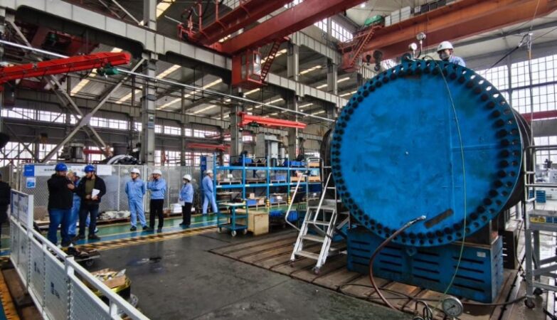 Uma visão detalhada e ampla do interior de uma movimentada fábrica industrial de fabricação pesada. À direita, destaca-se um componente industrial maciço, cilíndrico e azul brilhante, com uma enorme flange repleta de dezenas de parafusos, posicionado em um suporte azul. Este equipamento robusto sugere a fabricação de componentes críticos para o setor de energias renováveis, como partes para turbinas hidrelétricas ou eólicas de grande porte. Um trabalhador com capacete branco e macacão azul está no topo do equipamento, ajustando conexões perto de manômetros e mangueiras. Uma escada de metal se apoia na base da estrutura. À esquerda, um grupo de oito pessoas, incluindo engenheiros e inspetores vestindo capacetes azuis e brancos e casacos azuis, está reunido no piso de concreto, alguns observando pranchetas ou o equipamento. O vasto espaço da oficina é caracterizado por sua estrutura metálica, guindastes de pórtico vermelhos suspensos no teto alto, grandes janelas que deixam entrar luz natural e várias outras máquinas e prateleiras de estoque ao fundo. Placas informativas e cercas de segurança também são visíveis.