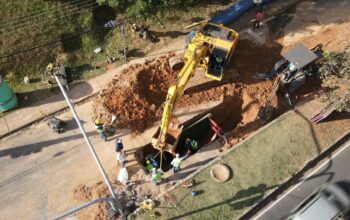 Vista aérea de um canteiro de obras em uma rua, onde maquinário pesado trabalha para instalar uma adutora para o transporte de água. Uma grande escavadeira amarela está posicionada na borda de uma vala profunda, removendo terra e depositando-a em uma pilha ao lado. Vários trabalhadores da construção civil, usando capacetes de segurança amarelos e coletes de alta visibilidade, estão dentro e ao redor da vala, supervisionando os trabalhos e movimentando materiais. Um trator menor está estacionado na rua adjacente. Um grande tubo azul está visível próximo à área de escavação, pronto para ser colocado na vala e compor o sistema de distribuição de água. A rua tem asfalto cinza e é ladeada por um gramado verde e árvores ao fundo. Um banheiro químico portátil está visível no canto superior esquerdo.