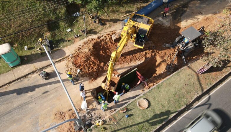 Vista aérea de um canteiro de obras em uma rua, onde maquinário pesado trabalha para instalar uma adutora para o transporte de água. Uma grande escavadeira amarela está posicionada na borda de uma vala profunda, removendo terra e depositando-a em uma pilha ao lado. Vários trabalhadores da construção civil, usando capacetes de segurança amarelos e coletes de alta visibilidade, estão dentro e ao redor da vala, supervisionando os trabalhos e movimentando materiais. Um trator menor está estacionado na rua adjacente. Um grande tubo azul está visível próximo à área de escavação, pronto para ser colocado na vala e compor o sistema de distribuição de água. A rua tem asfalto cinza e é ladeada por um gramado verde e árvores ao fundo. Um banheiro químico portátil está visível no canto superior esquerdo.