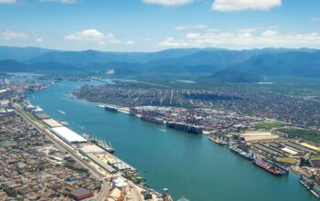 Uma vista aérea panorâmica do Porto de Santos, operado pela Autoridade Portuária de Santos (APS), com o canal estuarino central repleto de navios cargueiros e contêineres. À esquerda, a densa área urbana de Santos e, à direita, as instalações portuárias, armazéns e guindastes ladeando o Guarujá. Ao fundo, a cadeia de montanhas verdes sob um céu azul com nuvens brancas.