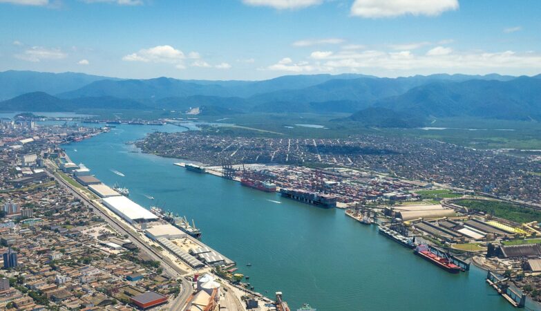 Uma vista aérea panorâmica do Porto de Santos, operado pela Autoridade Portuária de Santos (APS), com o canal estuarino central repleto de navios cargueiros e contêineres. À esquerda, a densa área urbana de Santos e, à direita, as instalações portuárias, armazéns e guindastes ladeando o Guarujá. Ao fundo, a cadeia de montanhas verdes sob um céu azul com nuvens brancas.