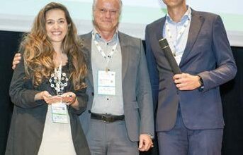Uma fotografia de premiação num palco, mostrando (da esquerda para a direita) Maria Gabriela da Climb Consulting, segurando um troféu transparente com detalhes prateados, ao lado de Sergei Fortes do Sinaenco, que está no centro com o braço no ombro dela, e Bernardo Etges da Climb Consulting, segurando um rolo de certificado e sorrindo. Os três profissionais estão formalmente vestidos, usando crachás de identificação e posando para a câmera.