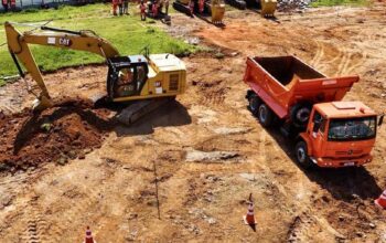 Vista aérea detalhada de um canteiro de obras ativo e terraplenado, representando as obras de extensão da Linha 4-Amarela. No primeiro plano, uma escavadeira hidráulica CAT amarela opera ativamente, cavando a terra marrom com sua concha. À sua direita, um caminhão caçamba laranja Mercedes-Benz está estacionado. Cones de segurança listrados estão posicionados na área de terra batida. Ao fundo, um grupo de operários com uniformes de segurança laranja está reunido perto de uma cerca branca que delimita o local, ao lado de outras duas escavadeiras amarelas estacionadas e de uma pequena pilha de pedras. A área é cercada por um muro de contenção e vegetação ao longe, sob luz solar direta.
