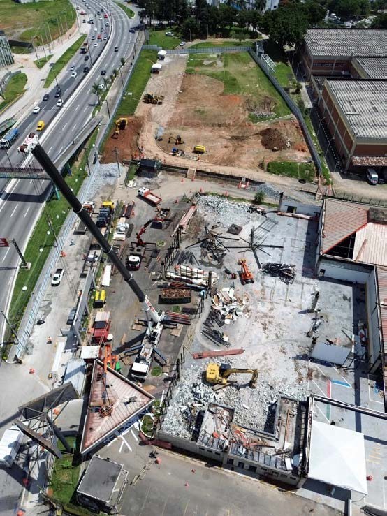 Uma vista aérea de um extenso canteiro de obras  da Linha 4-Amarela, em Taboão. A imagem mostra os estágios iniciais de demolição e preparação de terreno para um grande projeto de infraestrutura. No centro e primeiro plano, há entulho de concreto e estruturas parcialmente demolidas, com uma escavadeira amarela operando entre os detritos. Um guindaste alto está estendido e içando uma grande peça de treliça metálica. Vários caminhões e outros equipamentos de construção estão presentes. À esquerda, uma rodovia de várias faixas com tráfego intenso corre paralela ao local, com uma ponte pedonal a atravessá-la. Ao norte, uma grande área verde aberta com algumas árvores e tendas brancas menores faz fronteira com o canteiro. No lado direito, edifícios comerciais e industriais de concreto. A cena ilustra a atividade de renovação urbana em Taboão.