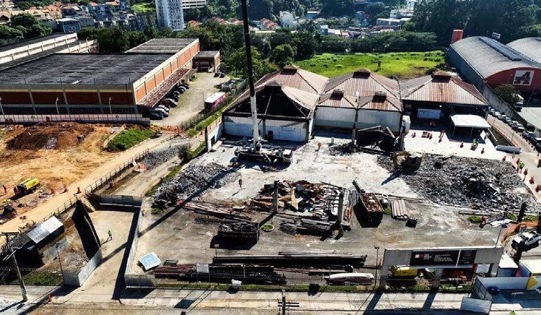 Uma vista aérea capturada em ângulo elevado mostra um canteiro de obras ativo e de demolição em uma área urbana de Taboão. No centro da imagem, montes de escombros, metal retorcido e detritos estão espalhados pelo chão de concreto, cercados por tapumes de segurança verdes. Um guindaste móvel branco com o braço estendido e uma escavadeira estão presentes no local. Atrás dos escombros, um complexo de edifícios antigos com telhados vermelhos em forma de pirâmide multi-emaranhados está parcialmente desmontado, adjacente a um galpão industrial moderno maior com telhado escuro. No primeiro plano inferior, há uma estrada e barreiras de segurança de concreto com longos tubos metálicos empilhados no chão. 