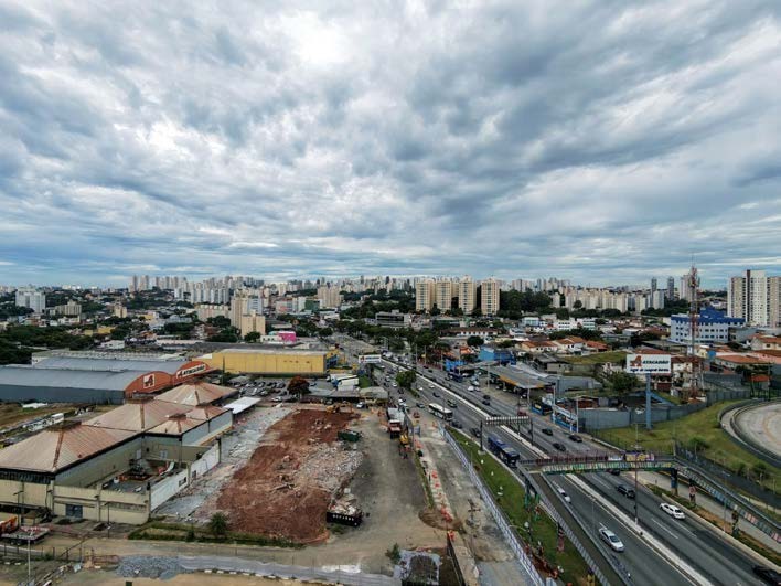 Uma vista aérea em ângulo elevado capturada da região de Taboão. No primeiro plano e centro-esquerda, destaca-se um amplo canteiro de obras de terraplanagem com terra vermelha exposta, adjacente a um galpão industrial mais antigo e a um grande galpão de metal laranja. À direita, uma via expressa de várias faixas corre verticalmente com tráfego de carros e ônibus, ao lado de um grande outdoor. O horizonte ao fundo é preenchido por um denso adensamento urbano de edifícios de apartamentos e escritórios, estendendo-se por toda a largura sob um céu pesadamente nublado e cinzento.