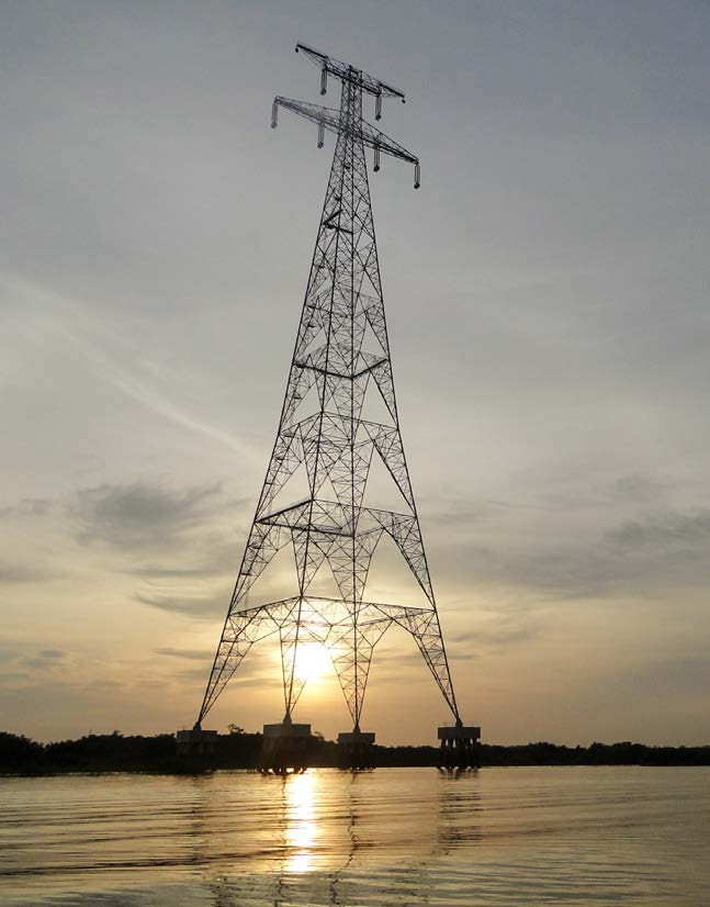 Uma fotografia de uma grande torre de transmissão de energia elétrica de treliça de aço, em pé em bases de concreto dentro de um corpo de água calmo ao pôr do sol. O sol baixo está posicionado perfeitamente atrás da base da torre, criando uma silhueta dramática e lançando um reflexo brilhante e ondulante na água em primeiro plano. O céu está nublado, com tons de cinza e laranja suave perto do horizonte. Margens arborizadas escuras são visíveis à distância. A torre de transmissão domina a composição central.