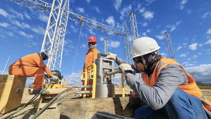 Fotografia de três trabalhadores de infraestrutura elétrica executando um trabalho de campo em um canteiro de obras de subestação. No primeiro plano, um homem agachado com capacete branco e colete laranja manipula uma ferramenta hidráulica em cabos. No fundo, dois outros trabalhadores em macacões laranja completo (um em pé de capacete vermelho e outro agachado de capacete branco) operam equipamentos hidráulicos e observam a operação. Ao fundo, estruturas metálicas de treliça em construção contra um céu azul com nuvens brancas.