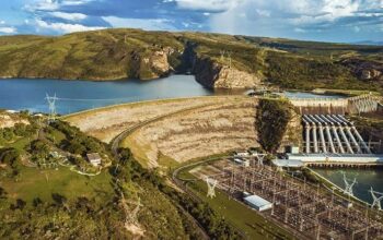 Uma vista aérea detalhada em grande angular de uma grande usina hidrelétrica situada em um vale cercado por colinas verdes e rochosas sob um céu parcialmente nublado. A imagem mostra uma barragem de arco de concreto substancial que retém um vasto reservatório de água azul. À direita da barragem, a casa de força é visível com grandes dutos de caneta metálicos alimentando o equipamento. No canto inferior direito, há uma grande subestação elétrica complexa preenchida com torres de metal, transformadores e fiação complexa. No primeiro plano e cruzando as colinas distantes, várias torres de transmissão de metal (treliças) com linhas de energia se estendem pela paisagem. O complexo inclui edifícios operacionais menores e estradas de acesso que se curvam ao redor das instalações. O reservatório azul calmo se estende profundamente entre as colinas rochosas, emoldurado pela paisagem natural.