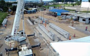 Nesta vista aérea detalhada de um canteiro de obras da Llucena Infraestrutura para o Túnel Serra Leste, vemos um grande guindaste branco posicionado no canto inferior esquerdo com estabilizadores estendidos sobre o solo arenoso. Seu braço estendido diagonalmente está içando uma robusta viga de concreto pré-moldado, um dos elementos fundamentais para a engenharia de pré-moldados do Túnel Serra Leste. Perto do centro da imagem, vários trabalhadores da Llucena Infraestrutura, usando capacetes e coletes refletivos, orientam e supervisionam o posicionamento da viga de pré-moldados ao lado de outra peça já instalada. O ambiente do canteiro mostra abrigos de lona azul, materiais de construção empilhados e uma fileira de ônibus azuis estacionados ao fundo, com a cidade e colinas no horizonte sob um céu nublado.