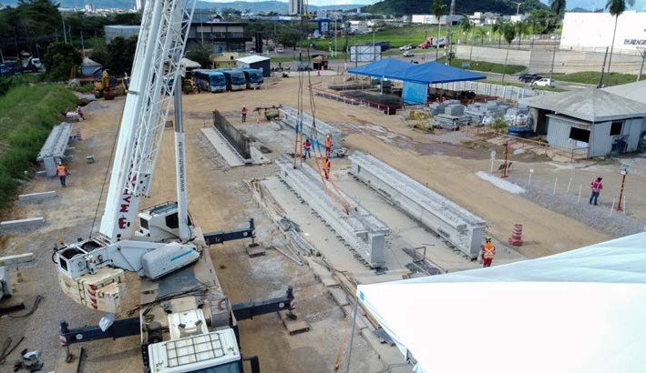 Nesta vista aérea detalhada de um canteiro de obras da Llucena Infraestrutura para o Túnel Serra Leste, vemos um grande guindaste branco posicionado no canto inferior esquerdo com estabilizadores estendidos sobre o solo arenoso. Seu braço estendido diagonalmente está içando uma robusta viga de concreto pré-moldado, um dos elementos fundamentais para a engenharia de pré-moldados do Túnel Serra Leste. Perto do centro da imagem, vários trabalhadores da Llucena Infraestrutura, usando capacetes e coletes refletivos, orientam e supervisionam o posicionamento da viga de pré-moldados ao lado de outra peça já instalada. O ambiente do canteiro mostra abrigos de lona azul, materiais de construção empilhados e uma fileira de ônibus azuis estacionados ao fundo, com a cidade e colinas no horizonte sob um céu nublado.