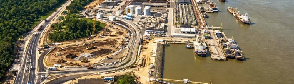 Uma vista aérea abrangente e detalhada de um complexo de infraestrutura multimodal em Porto Velho, Rondônia, à margem do Rio Madeira. A imagem mostra a integração entre uma moderna rodovia elevada (Nova 364) com tráfego de caminhões e os terminais portuários ativos no rio. No primeiro plano, o viaduto rodoviário contorna uma densa floresta Amazônica e passa por uma grande área de obra em andamento, incluindo terraplenagem, a construção de um túnel e rampas de acesso. Ao longo da margem do Rio Madeira, estendem-se terminais portuários de granéis com silos brancos, armazéns longos, guindastes e correias transportadoras. Vários navios de carga grandes e barcaças estão atracados nos píeres ou navegando no rio largo, ilustrando o escoamento de cargas em Porto Velho. A cidade de Porto Velho é visível ao longe na margem oposta do rio, sob um céu claro e ensolarado. A imagem enfatiza a expansão logística e a integração rodo-hidroviária na região.