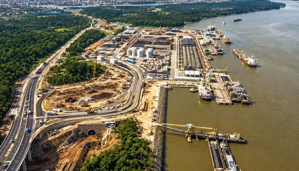 Uma vista aérea abrangente e detalhada de um complexo de infraestrutura multimodal em Porto Velho, Rondônia, à margem do Rio Madeira. A imagem mostra a integração entre uma moderna rodovia elevada (Nova 364) com tráfego de caminhões e os terminais portuários ativos no rio. No primeiro plano, o viaduto rodoviário contorna uma densa floresta Amazônica e passa por uma grande área de obra em andamento, incluindo terraplenagem, a construção de um túnel e rampas de acesso. Ao longo da margem do Rio Madeira, estendem-se terminais portuários de granéis com silos brancos, armazéns longos, guindastes e correias transportadoras. Vários navios de carga grandes e barcaças estão atracados nos píeres ou navegando no rio largo, ilustrando o escoamento de cargas em Porto Velho. A cidade de Porto Velho é visível ao longe na margem oposta do rio, sob um céu claro e ensolarado. A imagem enfatiza a expansão logística e a integração rodo-hidroviária na região.