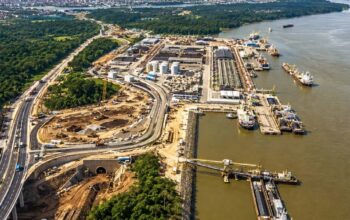 Uma vista aérea abrangente e detalhada de um complexo de infraestrutura multimodal em Porto Velho, Rondônia, à margem do Rio Madeira. A imagem mostra a integração entre uma moderna rodovia elevada (Nova 364) com tráfego de caminhões e os terminais portuários ativos no rio. No primeiro plano, o viaduto rodoviário contorna uma densa floresta Amazônica e passa por uma grande área de obra em andamento, incluindo terraplenagem, a construção de um túnel e rampas de acesso. Ao longo da margem do Rio Madeira, estendem-se terminais portuários de granéis com silos brancos, armazéns longos, guindastes e correias transportadoras. Vários navios de carga grandes e barcaças estão atracados nos píeres ou navegando no rio largo, ilustrando o escoamento de cargas em Porto Velho. A cidade de Porto Velho é visível ao longe na margem oposta do rio, sob um céu claro e ensolarado. A imagem enfatiza a expansão logística e a integração rodo-hidroviária na região.